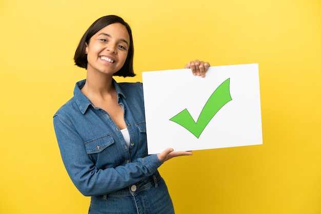 Mulher jovem de raça mista isolada em fundo amarelo segurando um cartaz com o texto Ícone de marca de seleção verde com expressão feliz