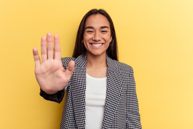 Mulher jovem de raça mista isolada em fundo amarelo, sorrindo alegre mostrando o número cinco com os dedos.