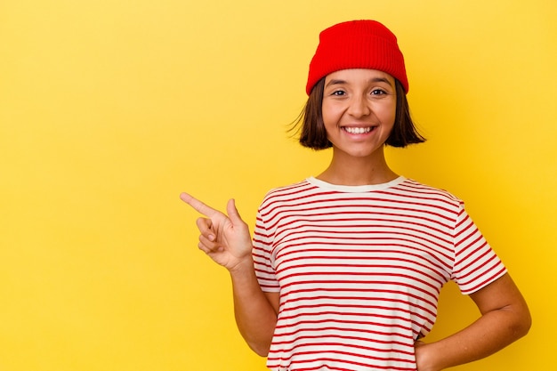 Mulher jovem de raça mista isolada em fundo amarelo, sorrindo alegremente, apontando com o dedo indicador para longe.