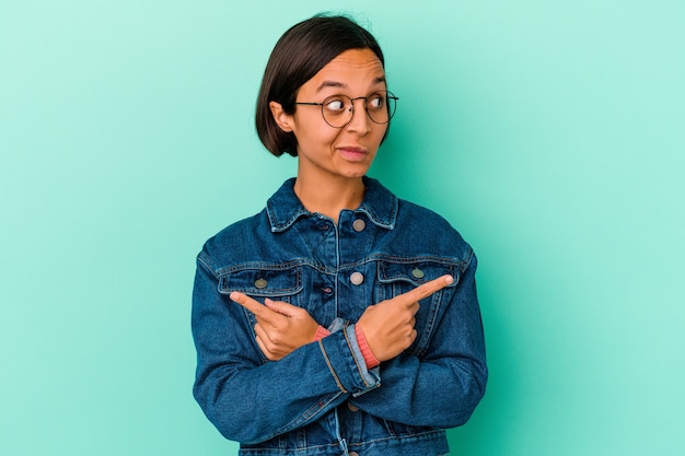 Mulher jovem de raça mista isolada em fundo azul aponta lateralmente, está tentando escolher entre duas opções.