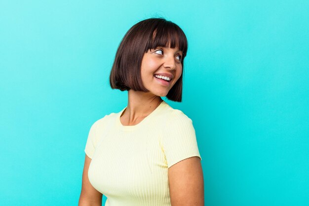 Mulher jovem de raça mista isolada em fundo azul parece de lado sorrindo, alegre e agradável.