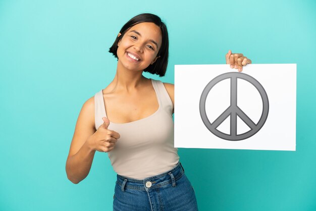 Foto mulher jovem de raça mista isolada em fundo azul segurando um cartaz com o símbolo da paz com o polegar para cima