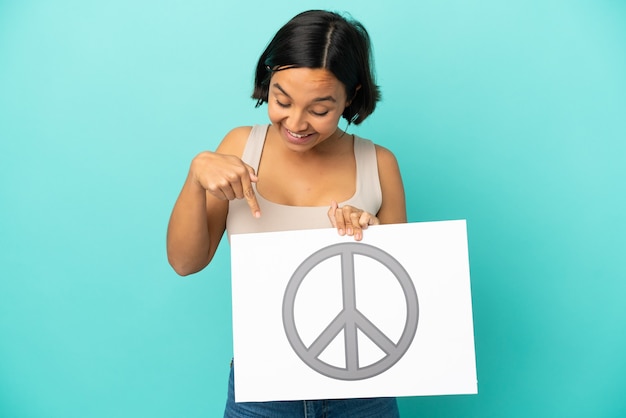 Foto mulher jovem de raça mista isolada em fundo azul segurando um cartaz com o símbolo da paz e apontando-o