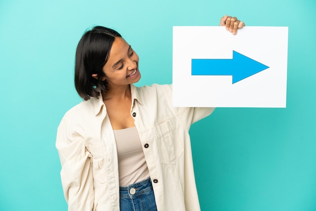 Mulher jovem de raça mista isolada em fundo azul segurando um cartaz com o símbolo de seta