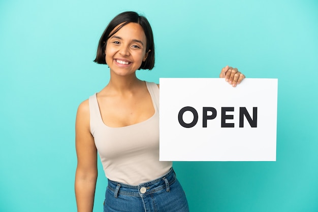 Mulher jovem de raça mista isolada em fundo azul segurando um cartaz com o texto ABERTO com expressão feliz