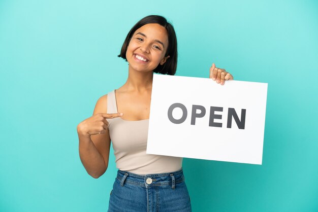 Mulher jovem de raça mista isolada em fundo azul segurando um cartaz com o texto ABRIR e apontando-o