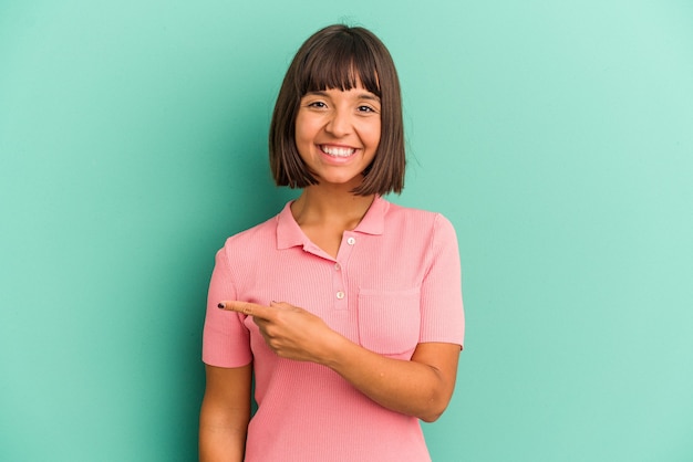 Mulher jovem de raça mista isolada em fundo azul, sorrindo e apontando de lado, mostrando algo no espaço em branco.