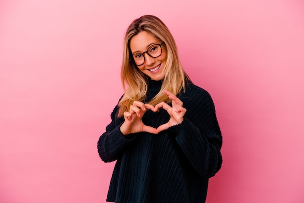 Mulher jovem de raça mista isolada em rosa sorrindo e mostrando uma forma de coração com as mãos.