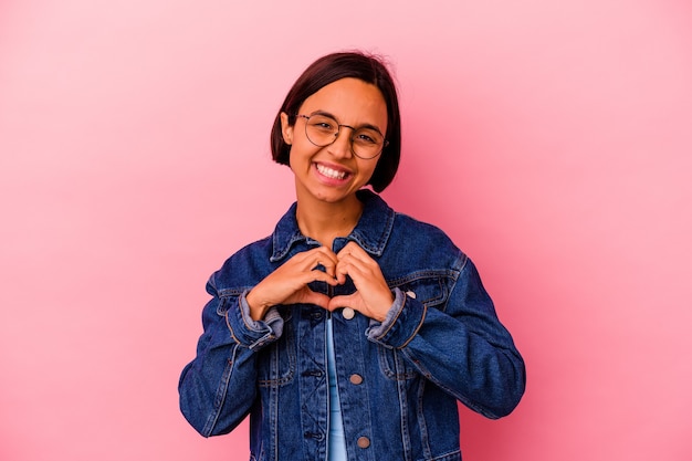 Mulher jovem de raça mista isolada na parede rosa, sorrindo e mostrando uma forma de coração com as mãos.