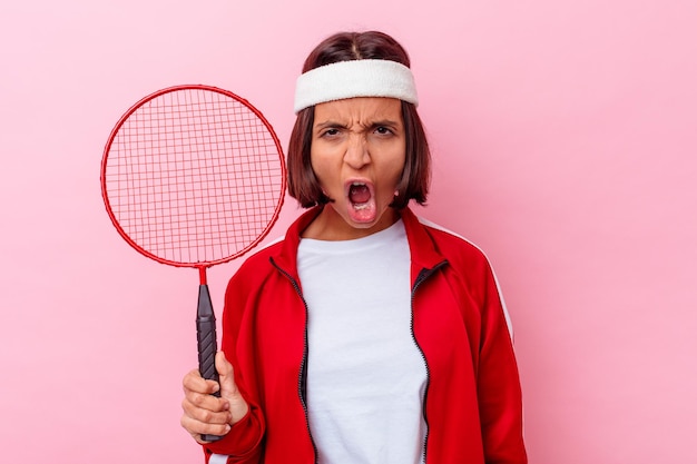 Mulher jovem de raça mista jogando badminton isolado na parede rosa, gritando muito zangada e agressiva.