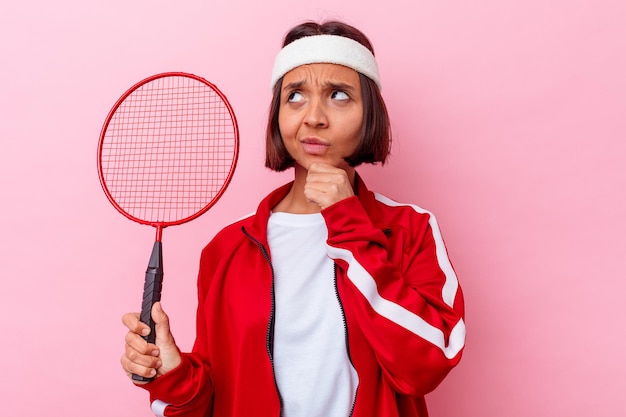 Mulher jovem de raça mista jogando badminton isolado na parede rosa, olhando de soslaio com expressão duvidosa e cética.