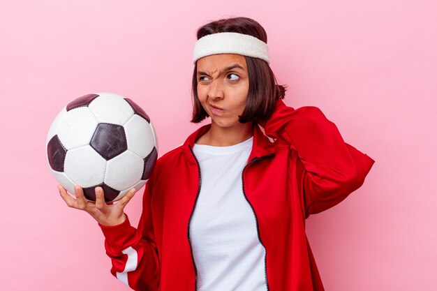 Mulher jovem de raça mista jogando futebol isolado na parede rosa, tocando a parte de trás da cabeça, pensando e fazendo uma escolha.