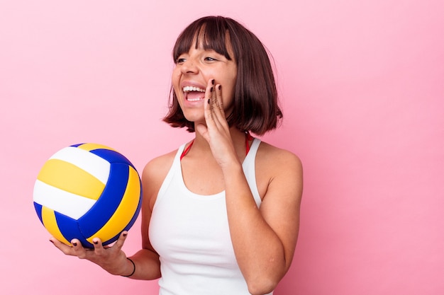 Mulher jovem de raça mista jogando vôlei isolado no fundo rosa, gritando e segurando a palma da mão perto da boca aberta.