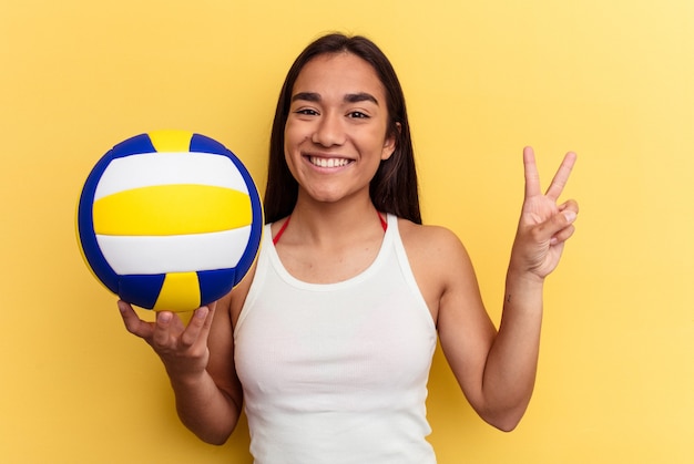 Mulher jovem de raça mista jogando vôlei na praia isolada em fundo amarelo alegre e despreocupada, mostrando um símbolo de paz com os dedos.