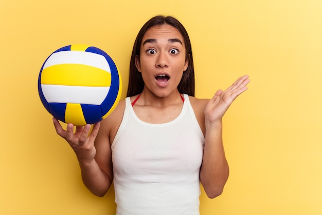 Mulher jovem de raça mista jogando vôlei na praia isolada em um fundo amarelo surpreso e chocado.