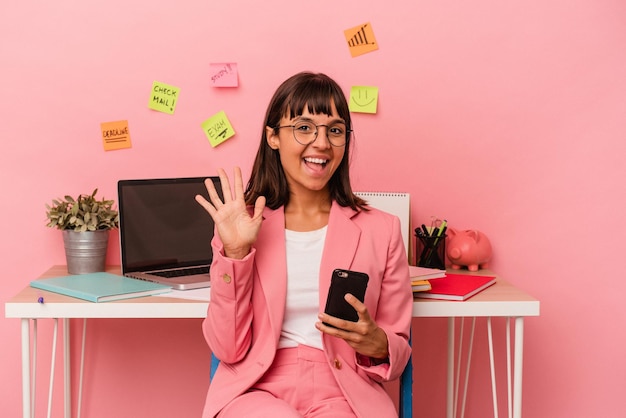 Mulher jovem de raça mista preparando um exame para a universidade segurando um celular isolado no fundo rosa sorrindo alegre mostrando o número cinco com os dedos