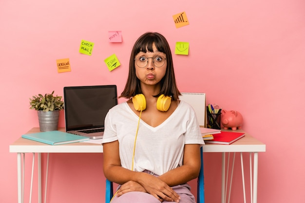 Mulher jovem de raça mista que prepara um exame na sala ouvindo música isolada no fundo rosa sopra nas bochechas, tem expressão cansada. Conceito de expressão facial.