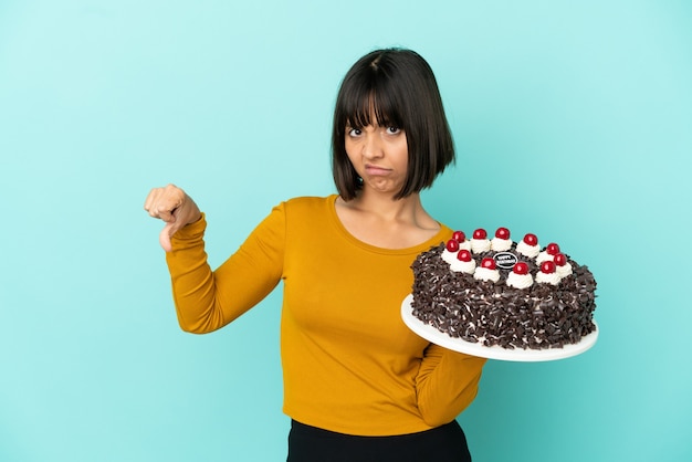 Mulher jovem de raça mista segurando bolo de aniversário e mostrando o polegar para baixo com expressão negativa