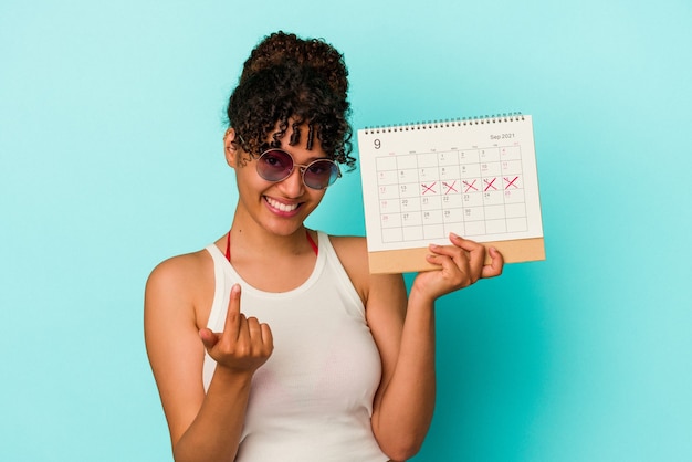 Mulher jovem de raça mista segurando calendário isolado em fundo azul, apontando com o dedo para você como se fosse um convite para se aproximar.