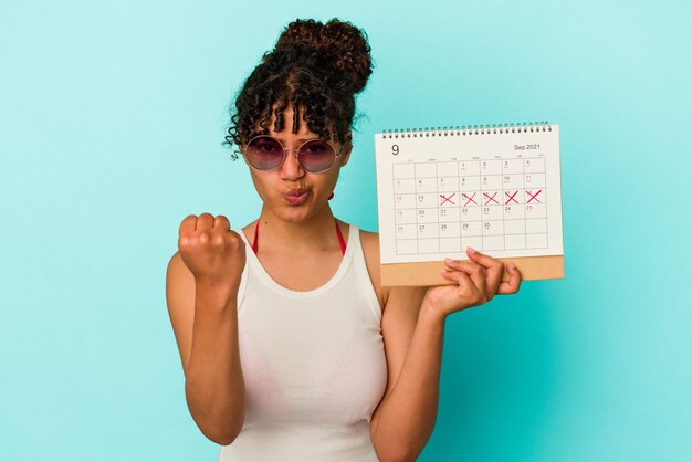 Mulher jovem de raça mista segurando calendário isolado em fundo azul, mostrando o punho para a câmera, expressão facial agressiva.