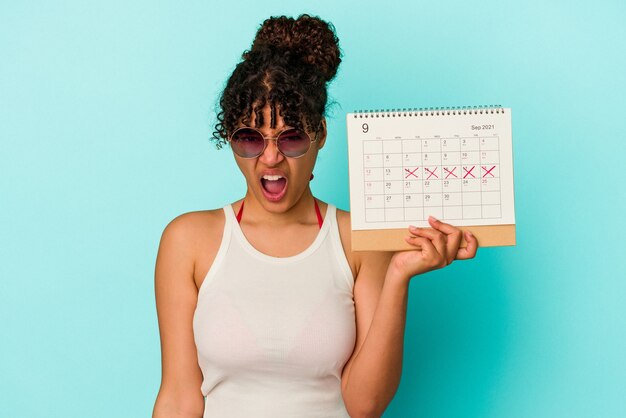 Foto mulher jovem de raça mista segurando calendário isolado no fundo azul, gritando muito zangado e agressivo.