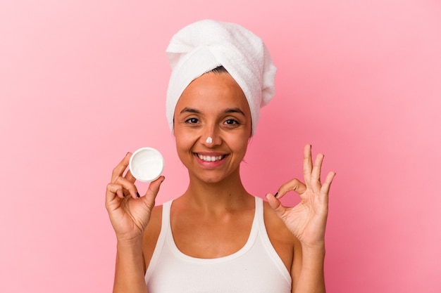 Mulher jovem de raça mista segurando creme facial isolado no fundo rosa alegre e confiante, mostrando um gesto ok.