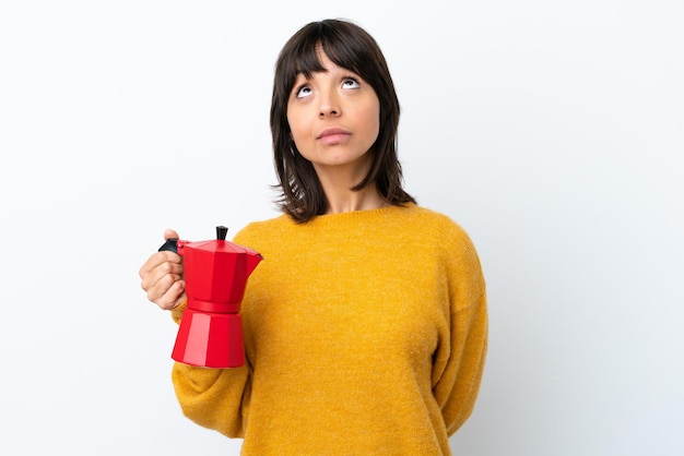 Mulher jovem de raça mista segurando o bule de café isolado no fundo branco e olhando para cima