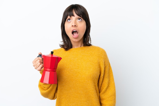 Mulher jovem de raça mista, segurando o bule de café isolado no fundo branco, olhando para cima e com expressão de surpresa