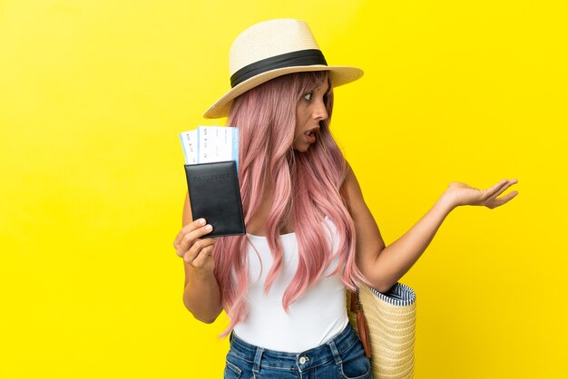 Mulher jovem de raça mista segurando passaporte e bolsa de praia isolados em um fundo amarelo com expressão de surpresa enquanto olha para o lado