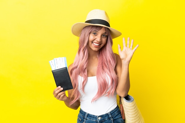 Mulher jovem de raça mista segurando passaporte e bolsa de praia isolados em um fundo amarelo e saudando com a mão com expressão feliz