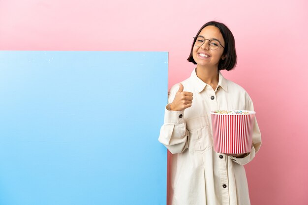 Mulher jovem de raça mista segurando pipocas com um grande banner sobre um fundo isolado fazendo um gesto de polegar para cima