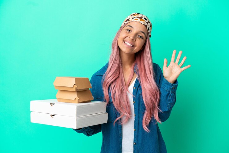Foto mulher jovem de raça mista segurando pizzas e hambúrgueres isolados no fundo verde e saudando com a mão com expressão feliz