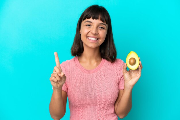 Mulher jovem de raça mista segurando um abacate isolado em fundo azul mostrando e levantando um dedo