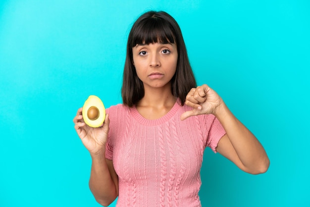 Mulher jovem de raça mista, segurando um abacate isolado em fundo azul, mostrando o polegar para baixo com expressão negativa