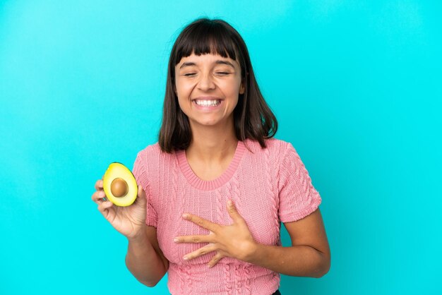 Mulher jovem de raça mista segurando um abacate isolado em um fundo azul e sorrindo muito