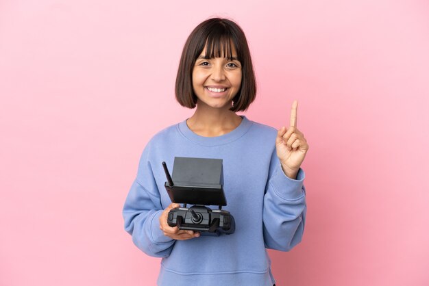 Mulher jovem de raça mista segurando um controle remoto de drone isolado em um fundo rosa, mostrando e levantando um dedo em sinal dos melhores