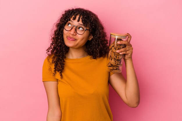 Mulher jovem de raça mista segurando um pote de biscoitos isolado em um fundo rosa e sonhando em alcançar objetivos e propósitos