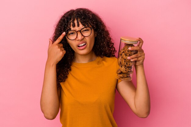 Mulher jovem de raça mista segurando um pote de biscoitos isolado no fundo rosa, mostrando um gesto de decepção com o dedo indicador.