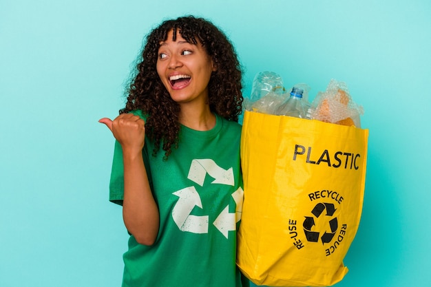 Mulher jovem de raça mista segurando um saco plástico reciclado isolado na parede azul aponta com o dedo polegar afastado, rindo e despreocupada.