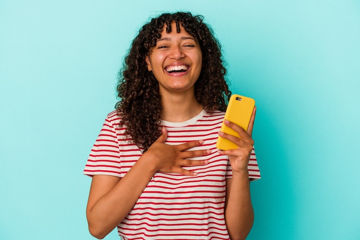 Foto mulher jovem de raça mista segurando um telefone móvel isolado sobre fundo azul ri alto, mantendo a mão no peito.