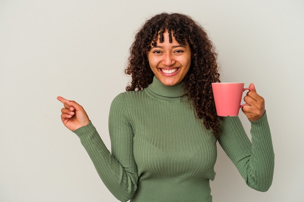 Mulher jovem de raça mista segurando uma caneca isolada no fundo branco, sorrindo e apontando para o lado, mostrando algo no espaço em branco.