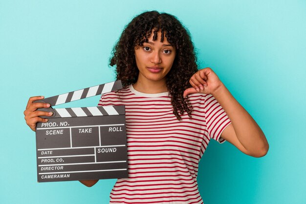 Mulher jovem de raça mista segurando uma claquete isolada sobre fundo azul, mostrando um gesto de antipatia, polegares para baixo. Conceito de desacordo.