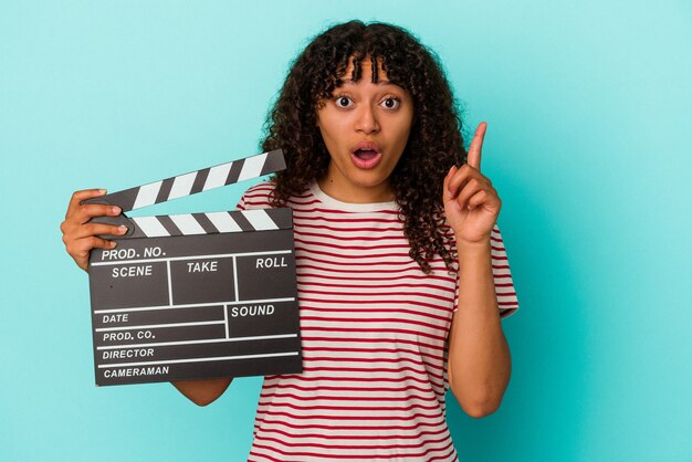 Mulher jovem de raça mista segurando uma claquete isolada sobre fundo azul, tendo uma ideia, o conceito de inspiração.