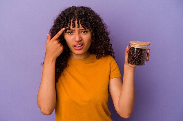 Mulher jovem de raça mista segurando uma garrafa de grãos de café isolada em um fundo roxo, mostrando um gesto de decepção com o dedo indicador.