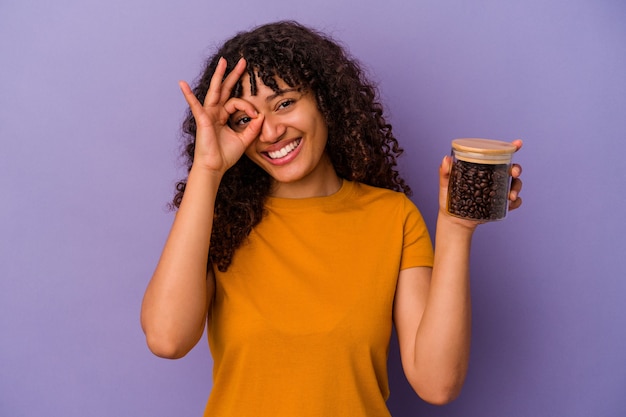 Mulher jovem de raça mista segurando uma garrafa de grãos de café isolada no fundo roxo animada mantendo o gesto ok no olho.