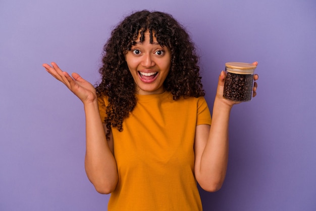 Mulher jovem de raça mista segurando uma garrafa de grãos de café isolada no fundo roxo, recebendo uma agradável surpresa, animada e levantando as mãos.