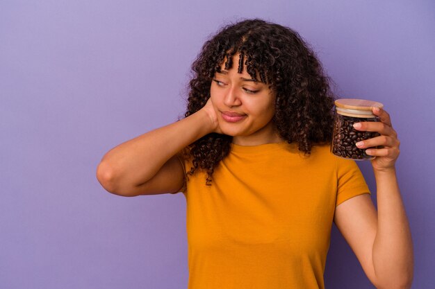 Mulher jovem de raça mista segurando uma garrafa de grãos de café isolada no fundo roxo tocando a parte de trás da cabeça, pensando e fazendo uma escolha.