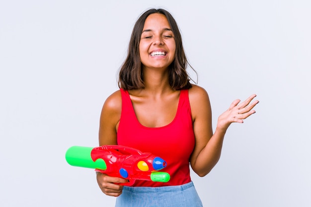 Mulher jovem de raça mista segurando uma pistola de água isolada ri alto, mantendo a mão no peito.