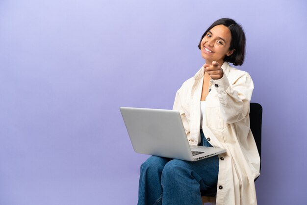 Mulher jovem de raça mista sentada em uma cadeira com laptop isolado em um fundo roxo apontando para a frente com uma expressão feliz