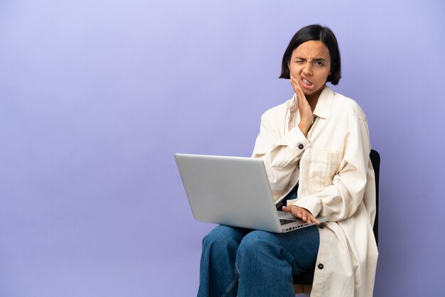 Mulher jovem de raça mista sentada em uma cadeira com laptop isolado em um fundo roxo com dor de dente